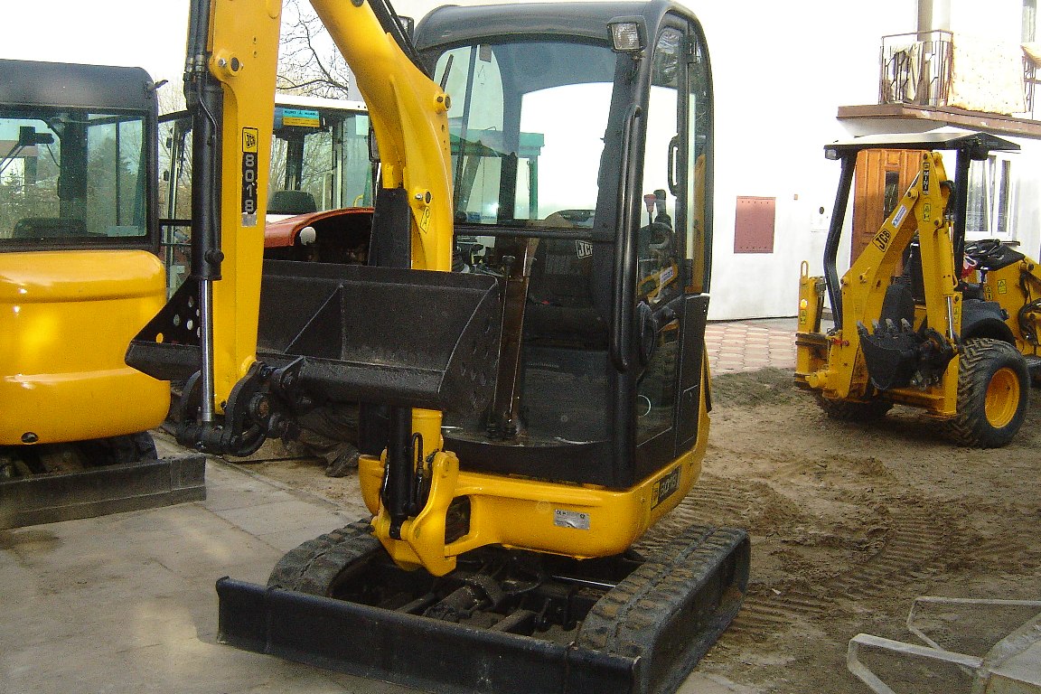 Żółta, kompaktowa maszyna budowlana JCB 8018 z rozsuwanymi gąsienicami, widoczna z przodu kabina, obok i w tle inne maszyny budowlane, na placu budowy.