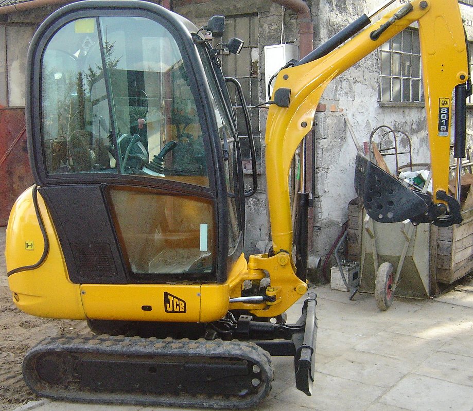 Żółta, gąsienicowa maszyna budowlana JCB 8018 z rozsuwanymi gąsienicami na tle budynku z cegły i wózka.