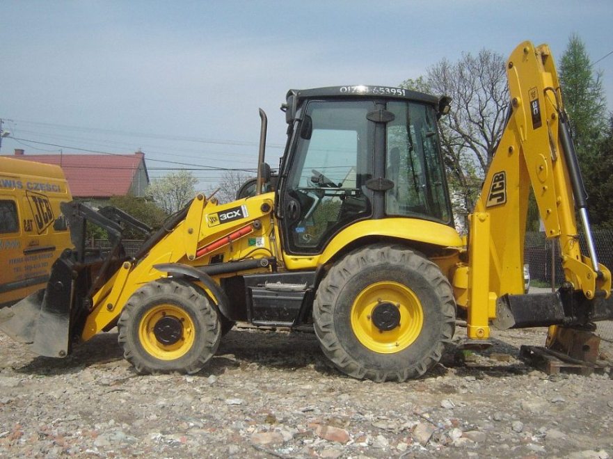 Żółta maszyna budowlana JCB 3CX Contractor z łyżką z przodu i ramieniem kopiącym z tyłu, stojąca na żwirowym terenie, w tle widoczny budynek mieszkalny i furgonetka serwisowa JCB.