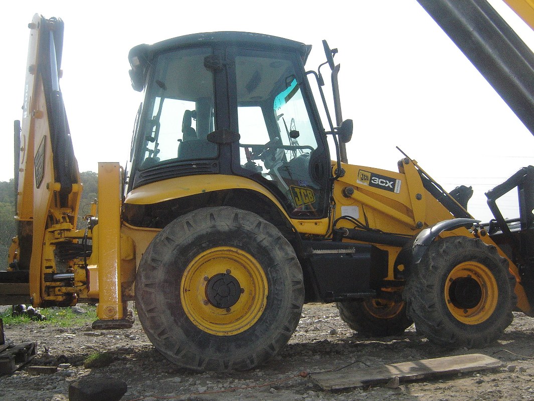 Żółta maszyna budowlana JCB 3CX Contractor z widocznymi oponami i kabiną operatora, stojąca na żwirowym podłożu.