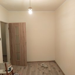 Vladyslav Pidhirnyi - Pomalowany pokój z drewnopodobnymi panelami na podłodze, otwartymi drzwiami z przeszkleniami, krzesłem z wiadrem farby i zwiniętym przedłużaczem elektrycznym przy ścianie.