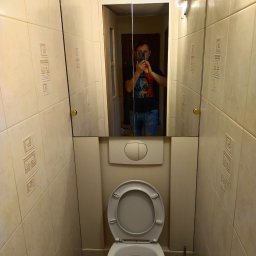 Vladyslav Pidhirnyi - Wąska toaleta wyłożona beżowymi płytkami z geometrycznym wzorem, widok na lustro z odbiciem osoby robiącej zdjęcie telefonem.