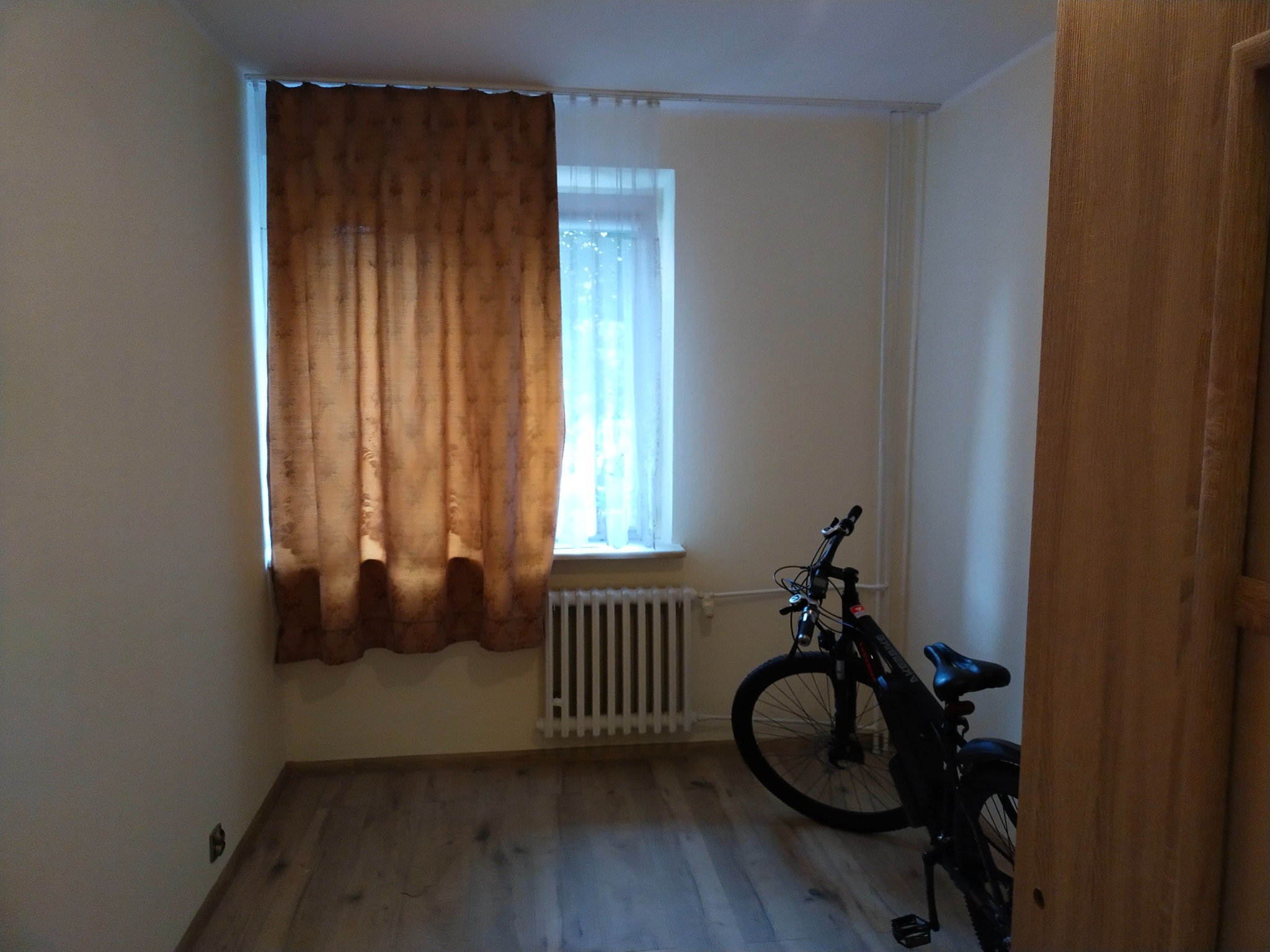 Pokój z drewnianą podłogą, zasłoniętym oknem i rowerem elektrycznym opartym o ścianę przy grzejniku.