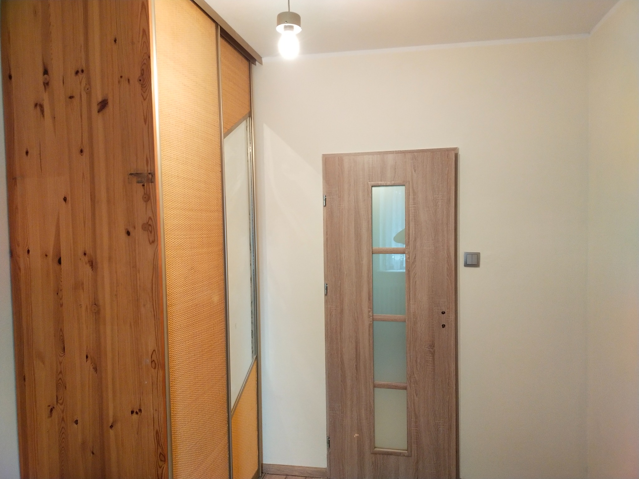 Wnętrze przedpokoju: drewniana szafa przesuwna z lustrem, drzwi wewnętrzne z przeszkleniami i włącznik światła na ścianie.