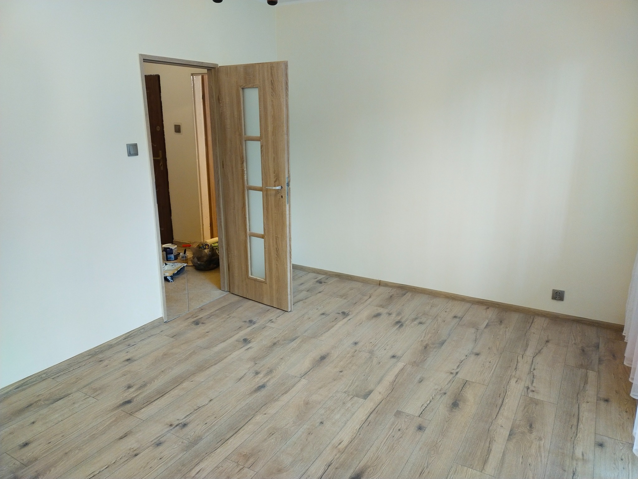 Jasne panele podłogowe ułożone w nowym pokoju z otwartymi drzwiami, widoczne ślady prac remontowych w sąsiednim pomieszczeniu.