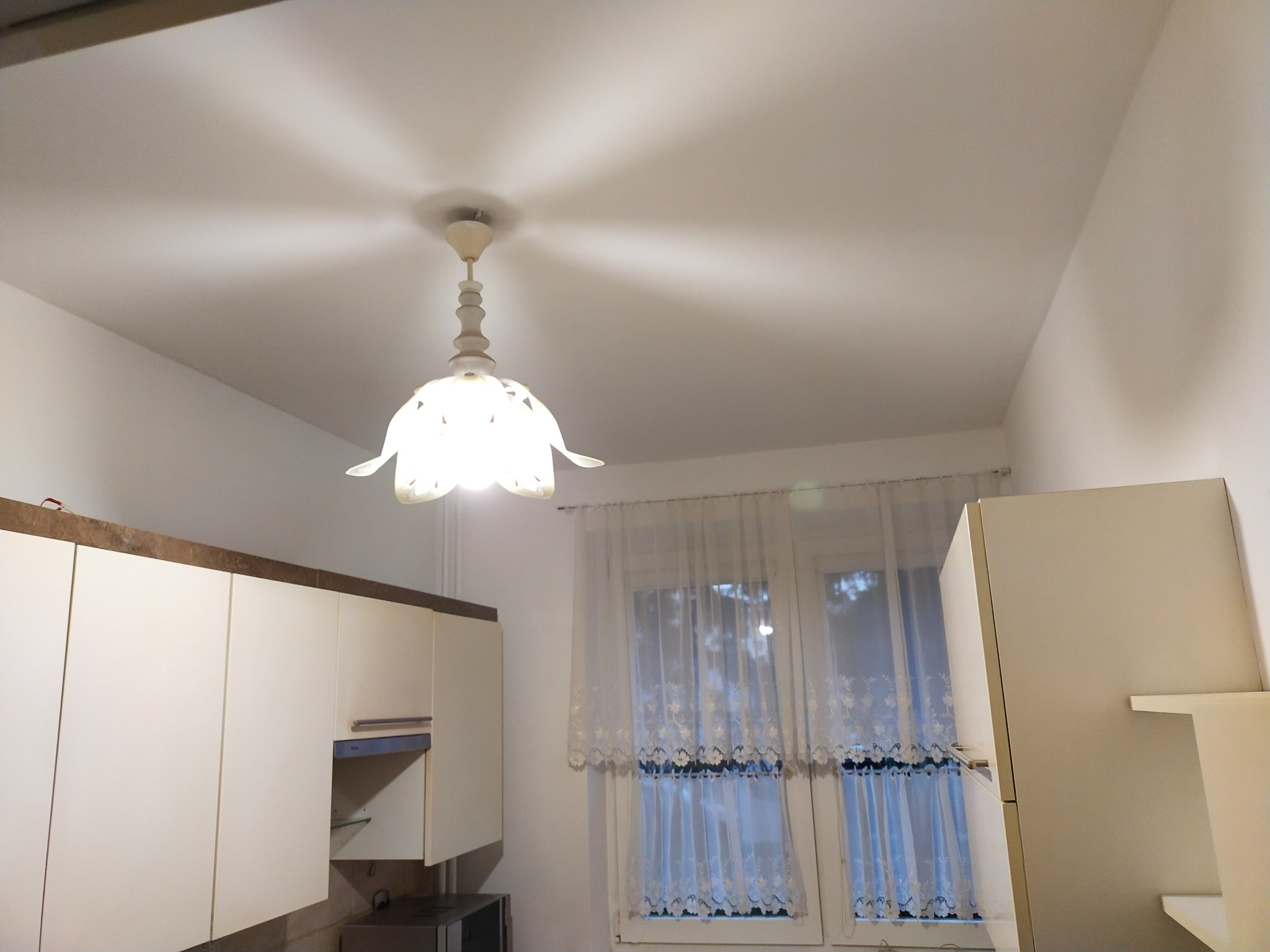 Kuchenne wnętrze z białymi szafkami, okno z firanką i lampą sufitową w kształcie kwiatu, widziane z dołu