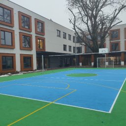 andrey karimov - Nowoczesny budynek z brązowymi ramami okiennymi widziany z boiska sportowego z niebiesko-zieloną nawierzchnią i bramką do piłki nożnej.