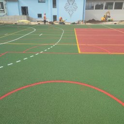 andrey karimov - Budowa boiska wielofunkcyjnego z zieloną i czerwoną nawierzchnią, widoczni pracownicy w pomarańczowych kamizelkach i koparka w tle.