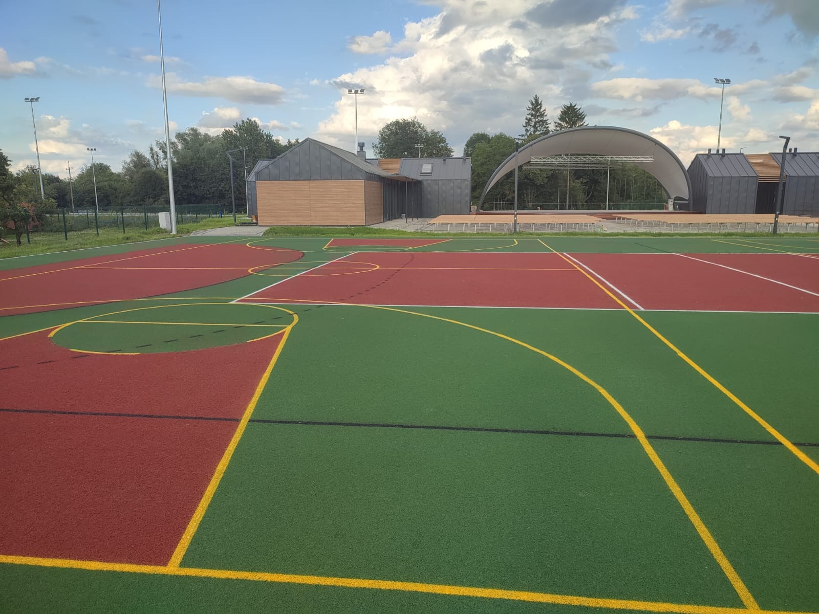 Wielofunkcyjne boisko sportowe z nawierzchnią tartanową w kolorach zieleni i czerwieni, z widocznymi liniami wyznaczającymi pola gry, na tle budynku z szarej blachy i drewnianą elewacją...