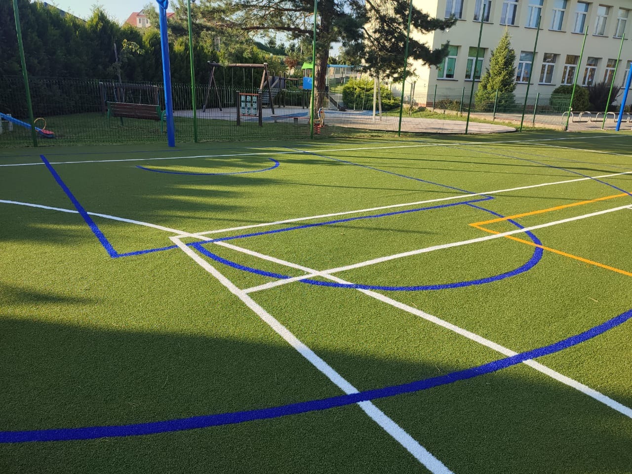 Nowo wybudowane boisko sportowe ze sztuczną trawą i wyraźnie oznaczonymi liniami w kolorze białym, niebieskim i pomarańczowym, widoczne elementy placu zabaw w tle.