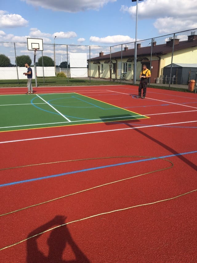 Malowanie linii na nowym, czerwono-zielonym boisku do koszykówki, z widocznymi zwojami sznurka i dwoma pracownikami w tle.