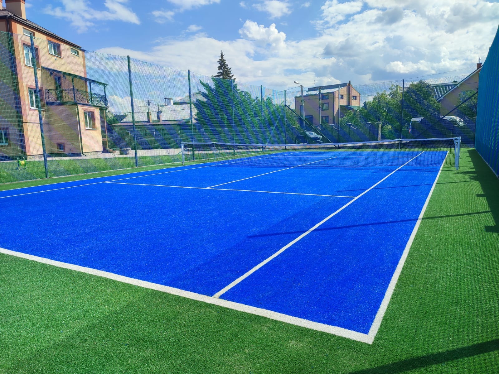 Niebieski kort tenisowy z zieloną sztuczną trawą wokół, otoczony siatką, w tle budynki i błękitne niebo z chmurami.