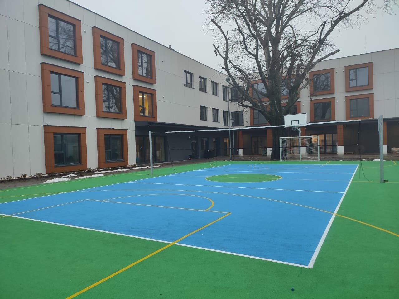 Nowoczesny budynek z brązowymi ramami okiennymi widziany z boiska sportowego z niebiesko-zieloną nawierzchnią i bramką do piłki nożnej.