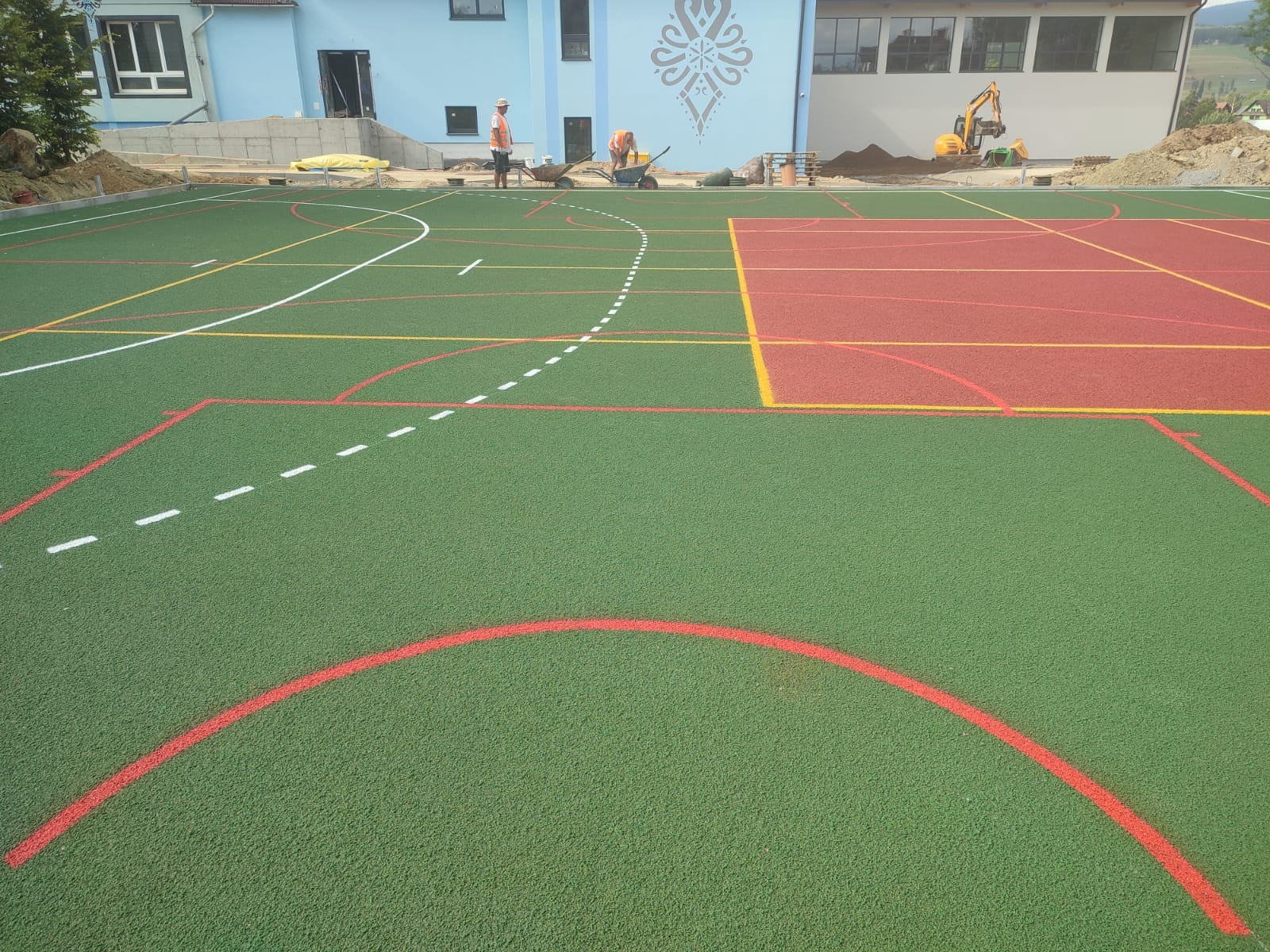 Budowa boiska wielofunkcyjnego z zieloną i czerwoną nawierzchnią, widoczni pracownicy w pomarańczowych kamizelkach i koparka w tle.