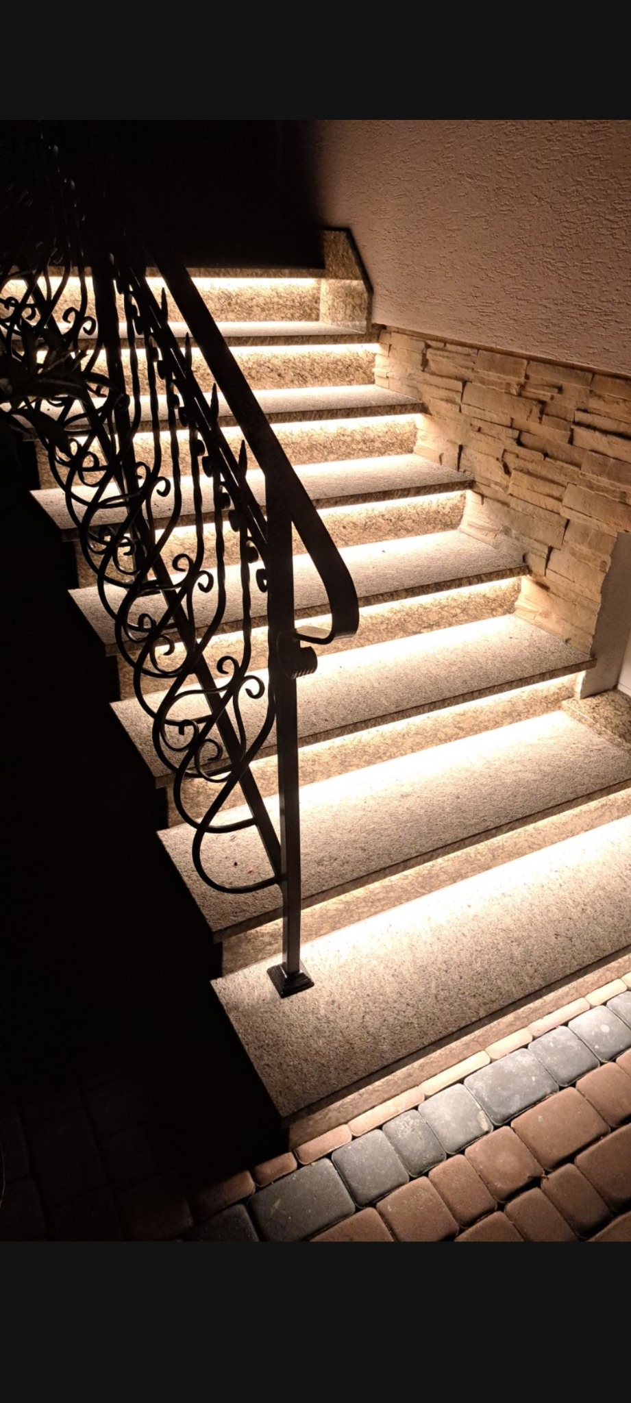 Podświetlane schody kamienne z kutą balustradą, oświetlenie LED podkreśla fakturę kamienia i dodaje elegancji. Widoczny fragment kamiennej ściany i kostki brukowej.