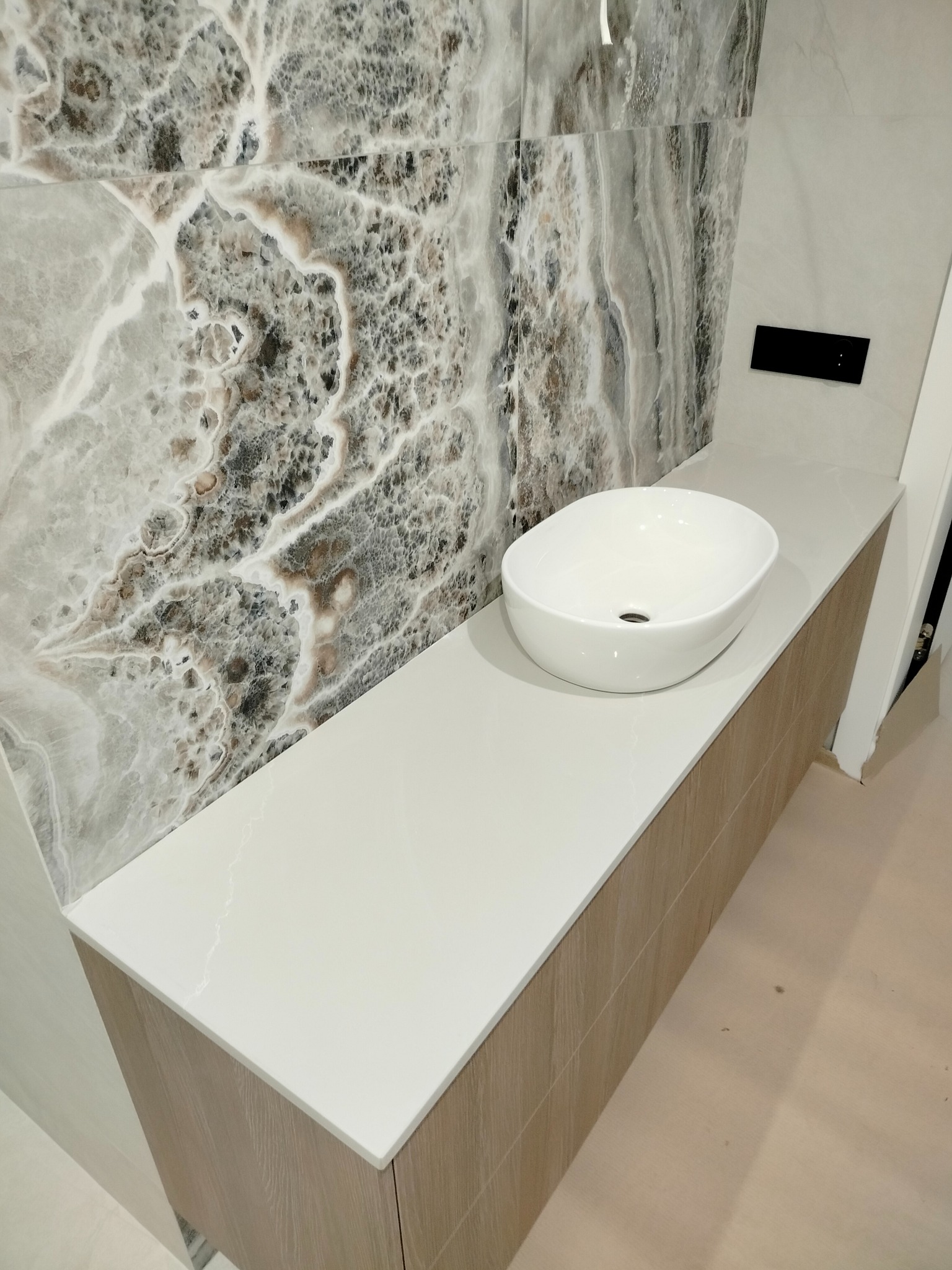 Biały blat kamienny z umywalką nablatową w łazience, na tle ściany z marmurowym wzorem. Ujęcie z perspektywy, ukazujące detale wykończenia i nowoczesny design.