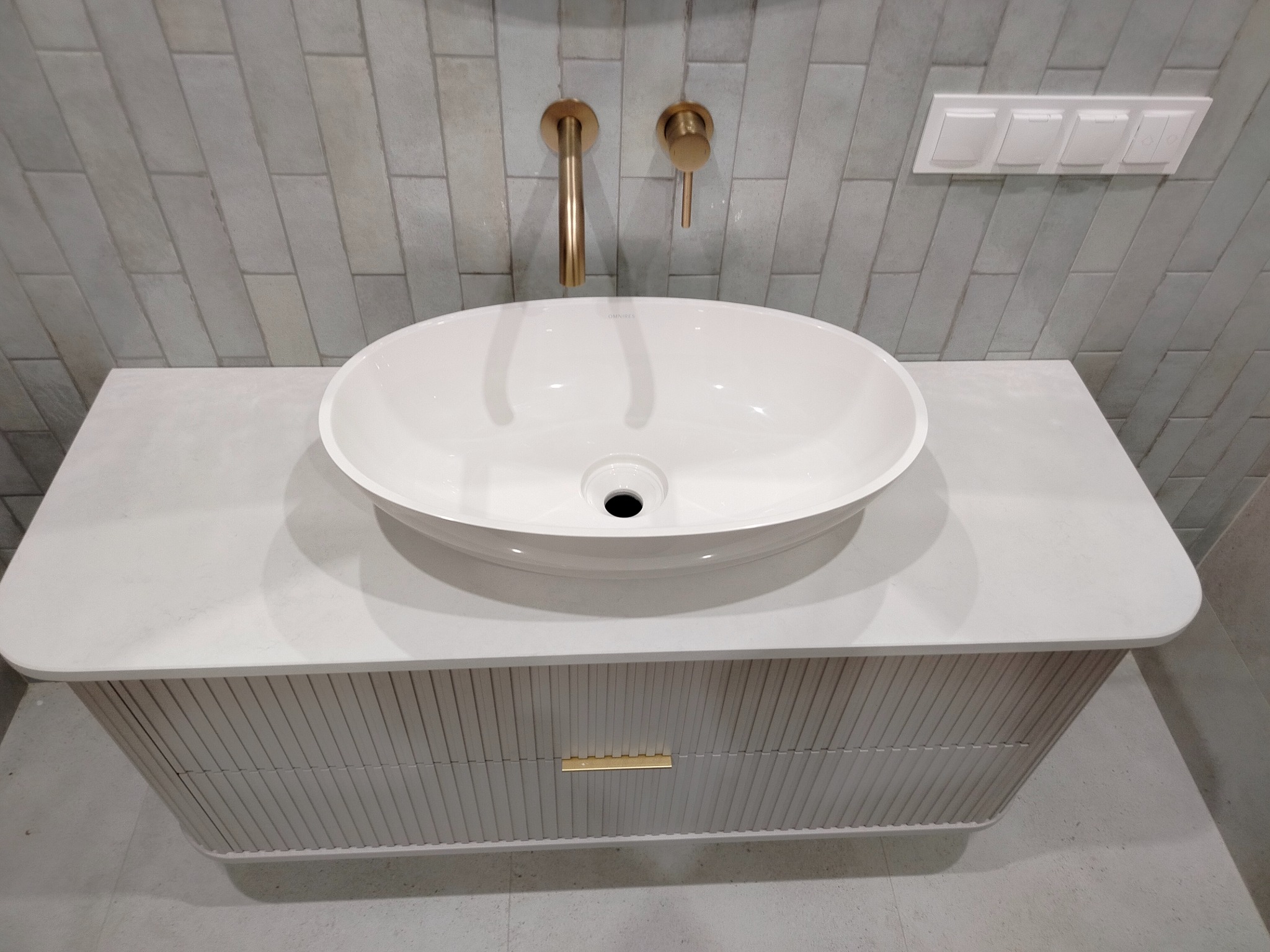 Biała, owalna umywalka nablatowa na blacie z konglomeratu, złota armatura ścienna, szare płytki na ścianie. Nowoczesna łazienka.