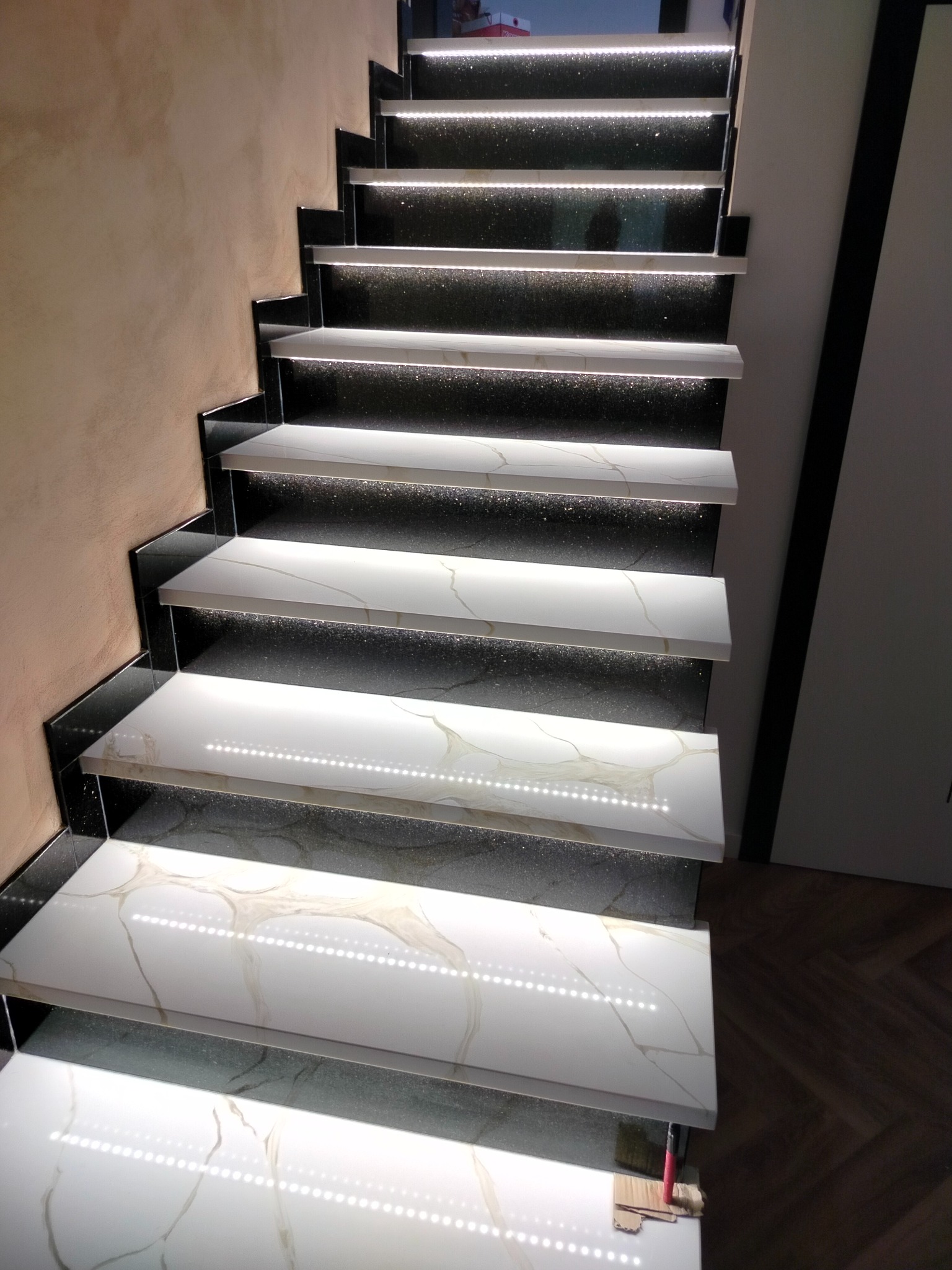 Nowoczesne schody z podświetleniem LED, białe stopnie z marmurowym wzorem i czarne boki. Widok z dołu, podkreślający elegancki design i precyzję wykonania.