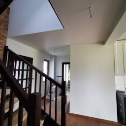 Mura-Dom - Wnętrze domu po remoncie: świeżo pomalowane ściany na biało, drewniane schody zabezpieczone taśmą malarską, widoczna ceglana ściana i fragment sufitu z wystającym kablem oświetleniowym.