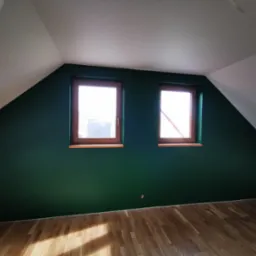 Poddasze po remoncie: ściana pomalowana na ciemnozielony kolor, drewniane okna i parkiet, białe skosy sufitu.