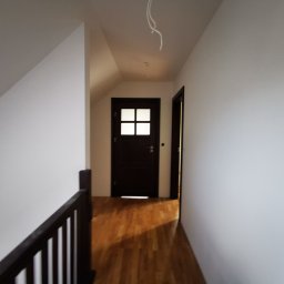 Mura-Dom - Korytarz na poddaszu z drewnianą podłogą, białymi ścianami i ciemnymi drzwiami z przeszkleniami, widoczne kable oświetleniowe wystające z sufitu.