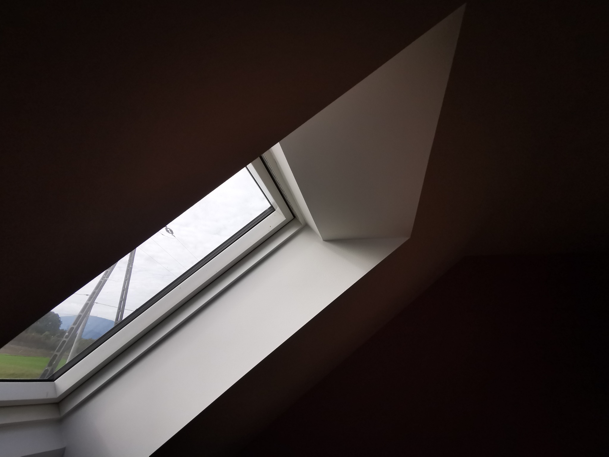 Okno dachowe z widokiem na zielone pola i słupy energetyczne, wnęka pomalowana na biało, kontrast z ciemnym sufitem poddasza.