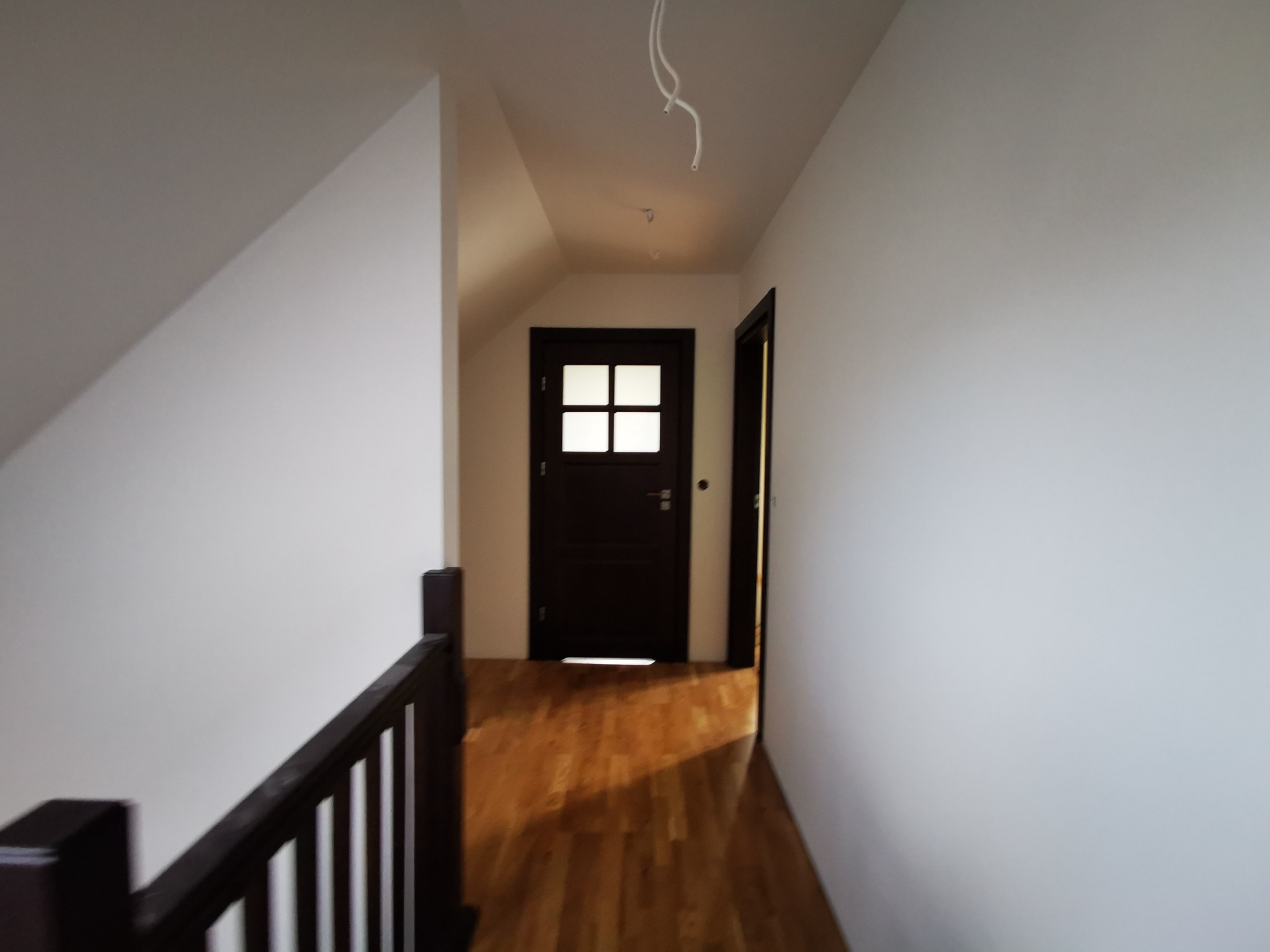 Korytarz na poddaszu z drewnianą podłogą, białymi ścianami i ciemnymi drzwiami z przeszkleniami, widoczne kable oświetleniowe wystające z sufitu.