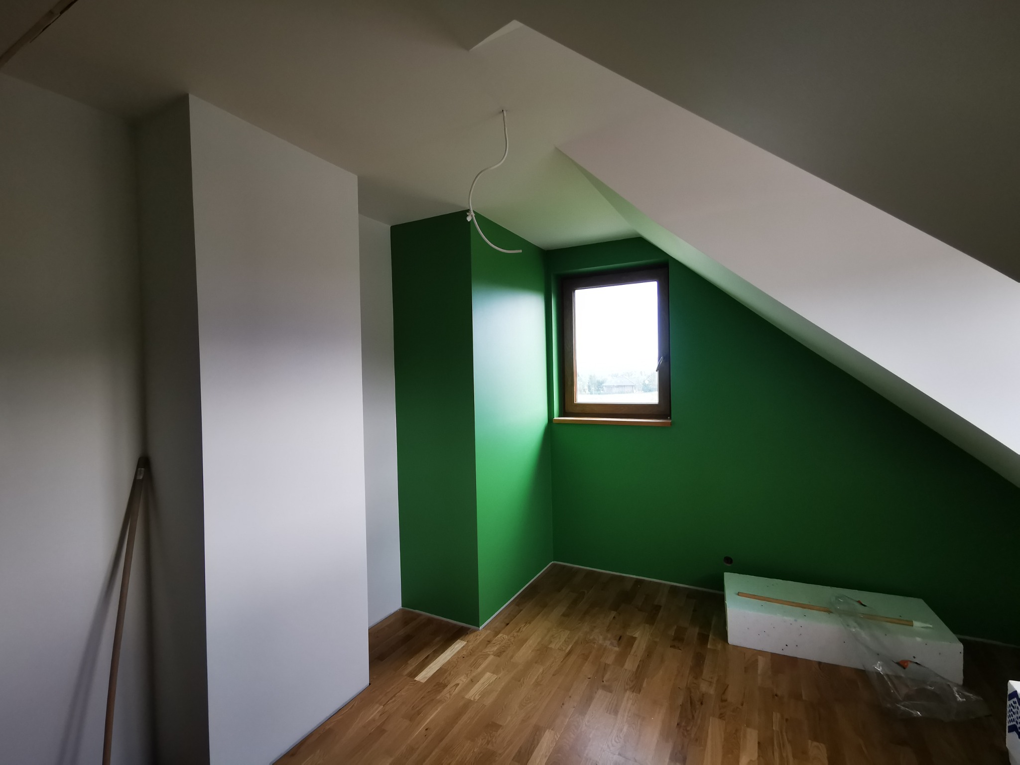 Poddasze po remoncie: zielone ściany, drewniane okno, białe skosy, parkiet i styropian, widoczne kable zwisające z sufitu.