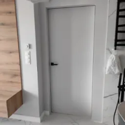 Nowoczesna łazienka z ukrytymi drzwiami w kolorze białym, marmurowa podłoga i ściany, drewniany element zabudowy, biały włącznik światła i czarny grzejnik drabinkowy.