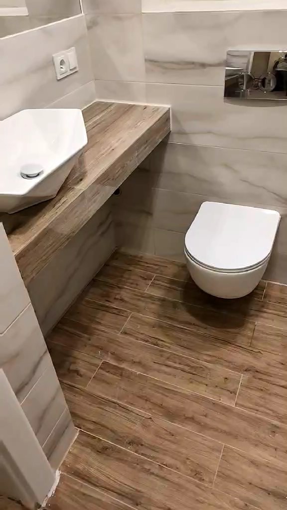 Nowoczesna łazienka z białymi płytkami imitującymi marmur, drewnopodobną podłogą z płytek, wiszącą miską WC i umywalką nablatową o geometrycznym kształcie na drewnianym blacie.