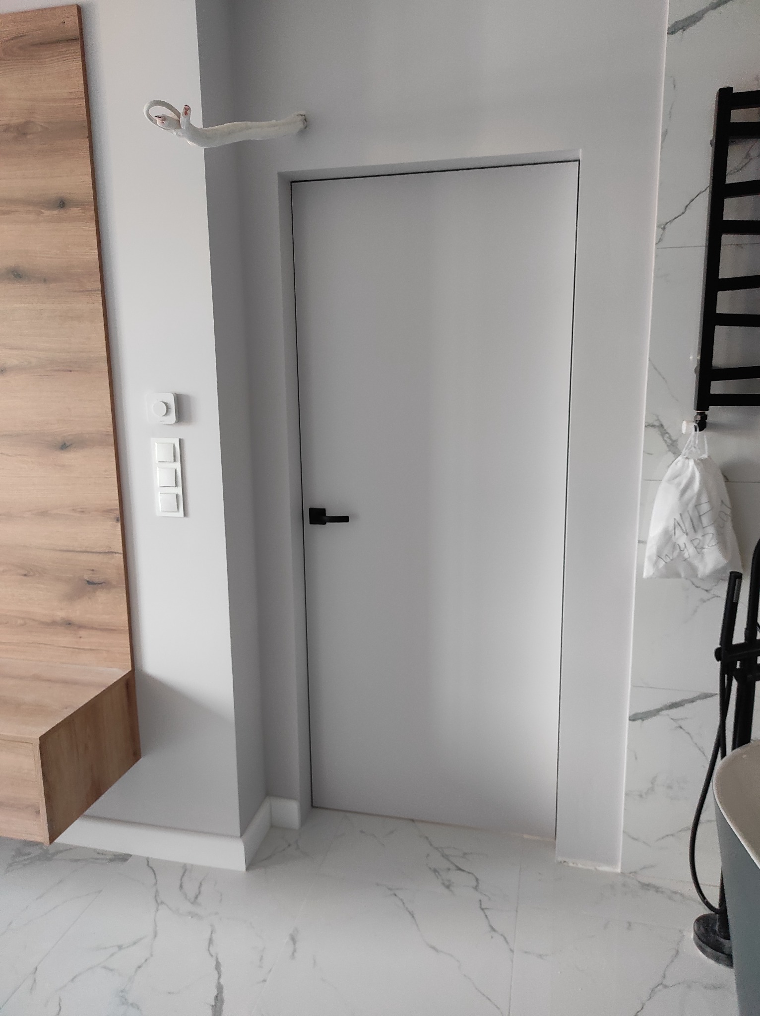 Nowoczesna łazienka z ukrytymi drzwiami w kolorze białym, marmurowa podłoga i ściany, drewniany element zabudowy, biały włącznik światła i czarny grzejnik drabinkowy.