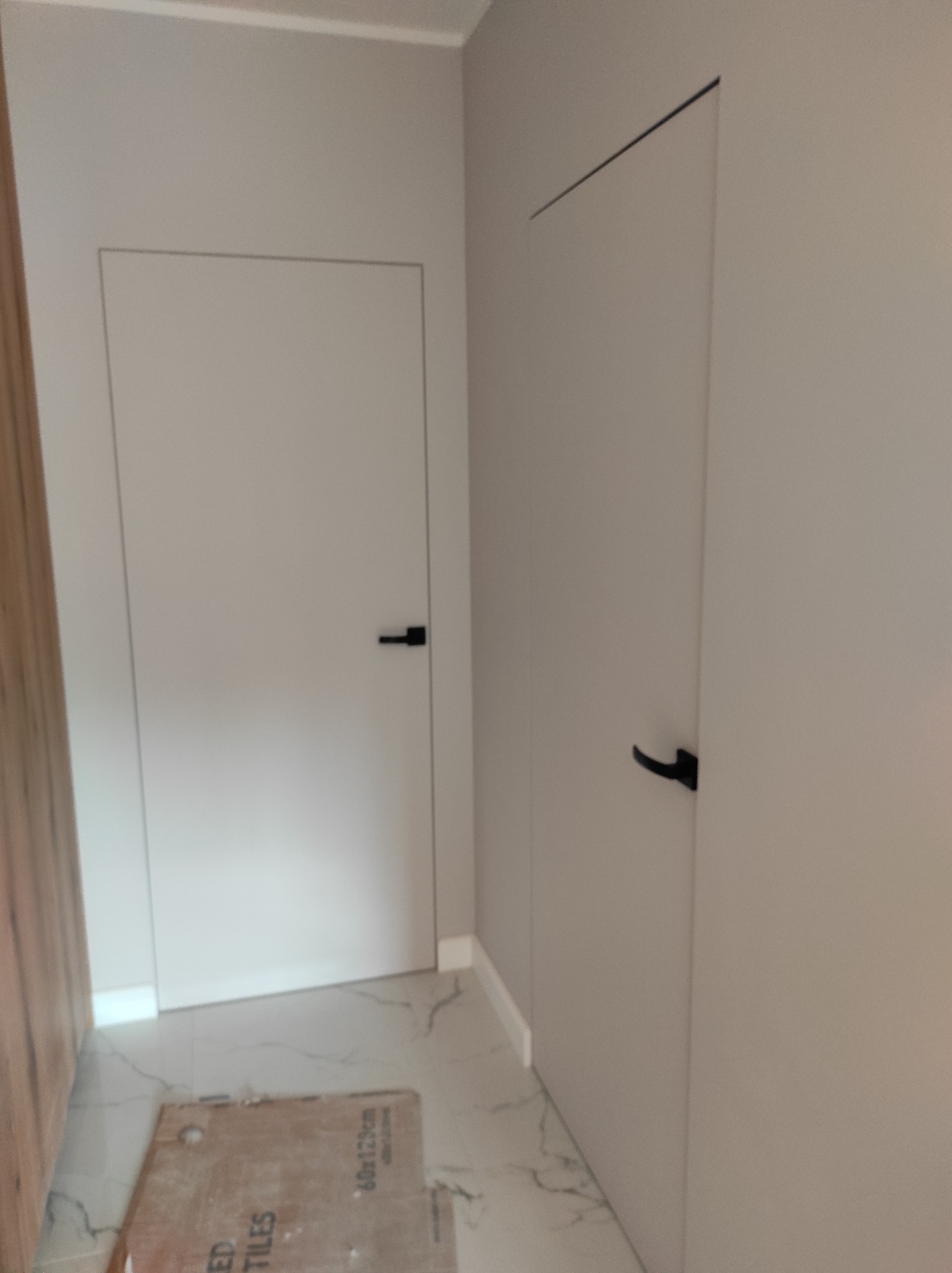 Dwa minimalistyczne, ukryte drzwi z czarnymi klamkami na tle jasnych ścian i marmurowej podłogi. Na podłodze leży karton po płytkach.