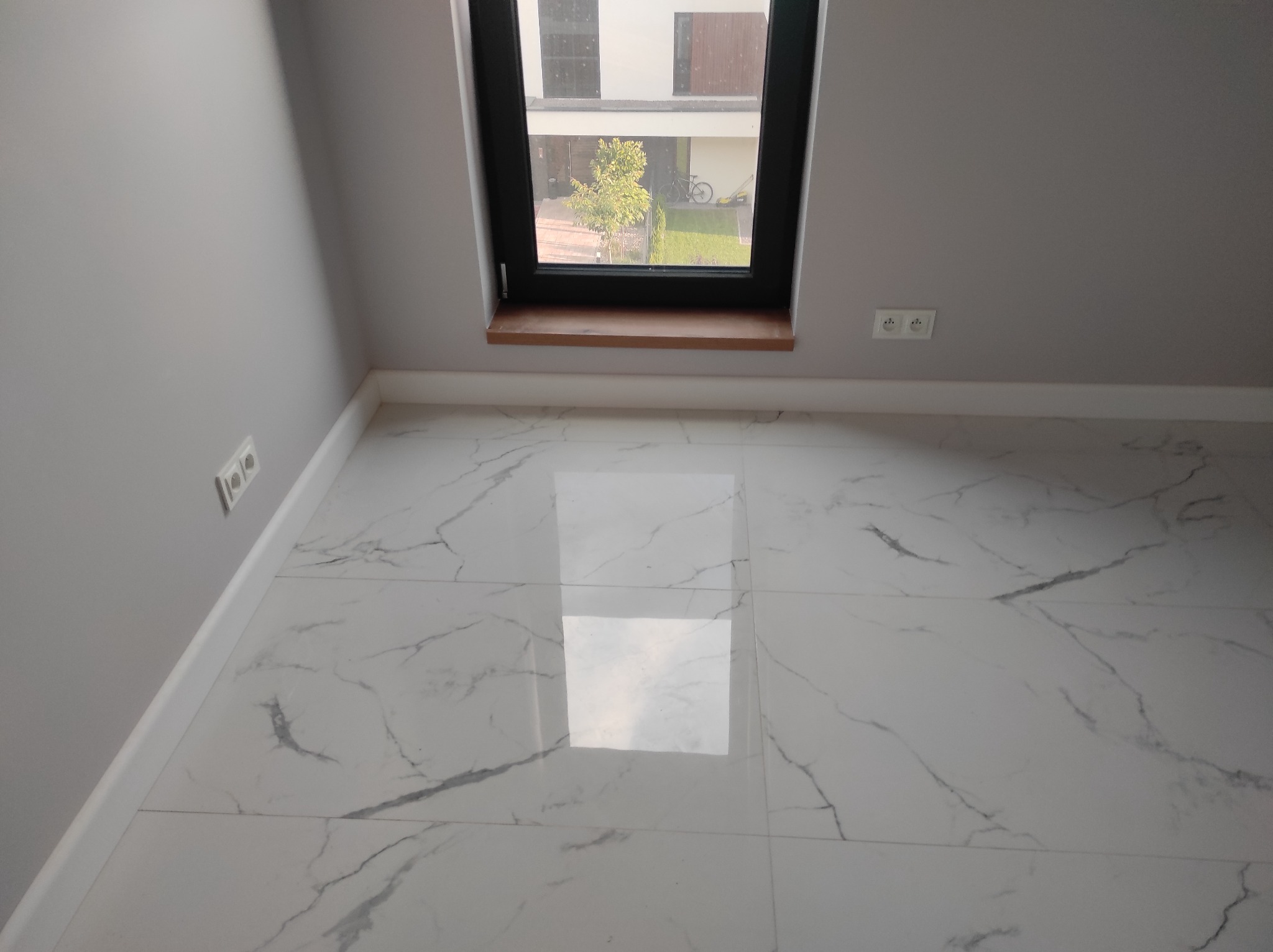 Wykończona podłoga z białych płytek imitujących marmur, widok na okno z drewnianym parapetem i fragment ściany z gniazdkami elektrycznymi.