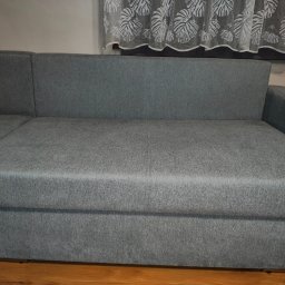 Daniel Stec - Szary narożnik z tkaniny w salonie, widok z przodu, z zasłoną w liście w tle. Widoczne drewniane panele podłogowe i fragment ściany. Minimalistyczny design.