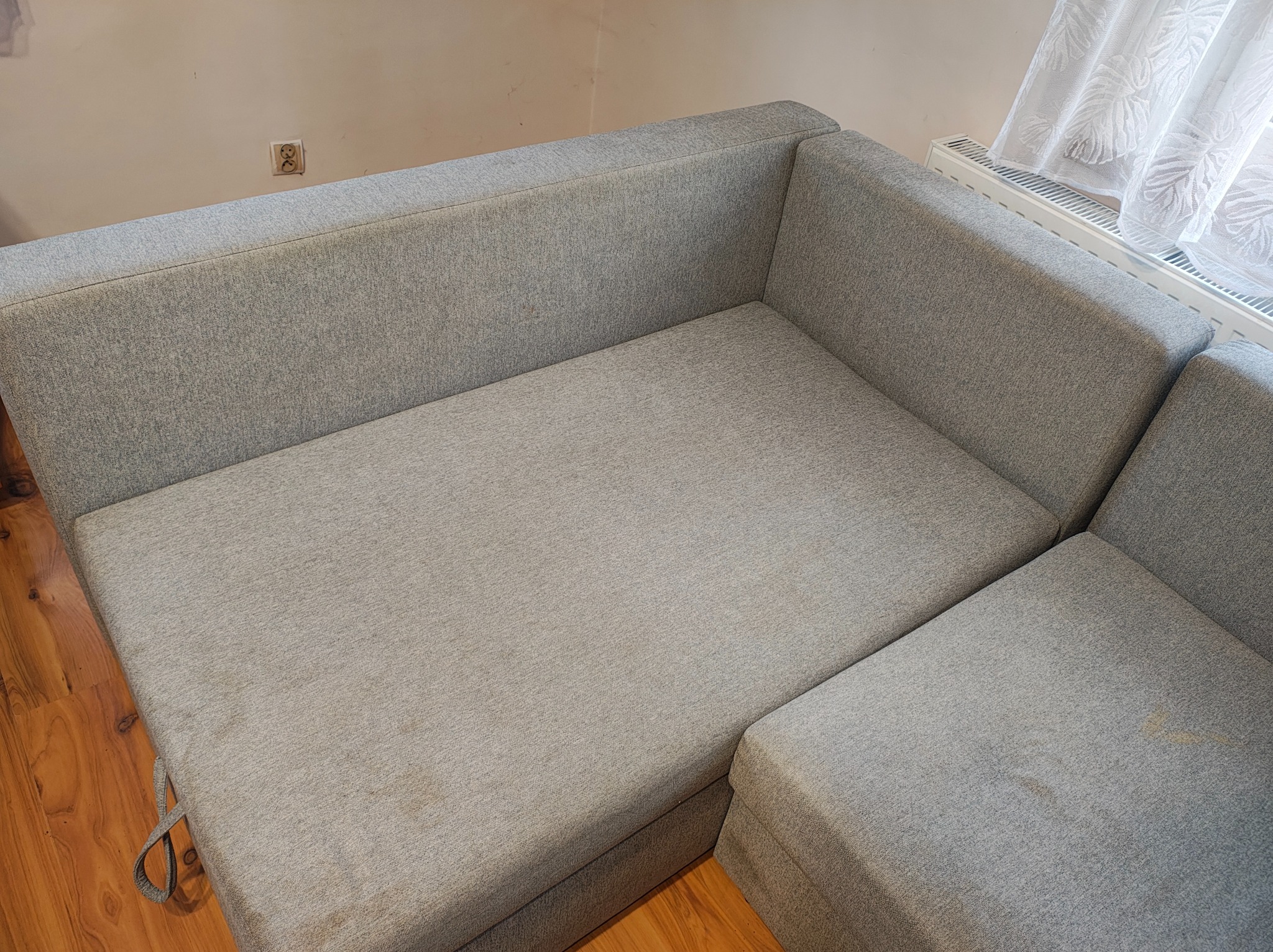 Szara, narożna kanapa z widocznymi plamami na siedzisku, ukazana z góry, z fragmentem drewnianej podłogi i zasłony w tle. Widoczne gniazdko elektryczne na ścianie.