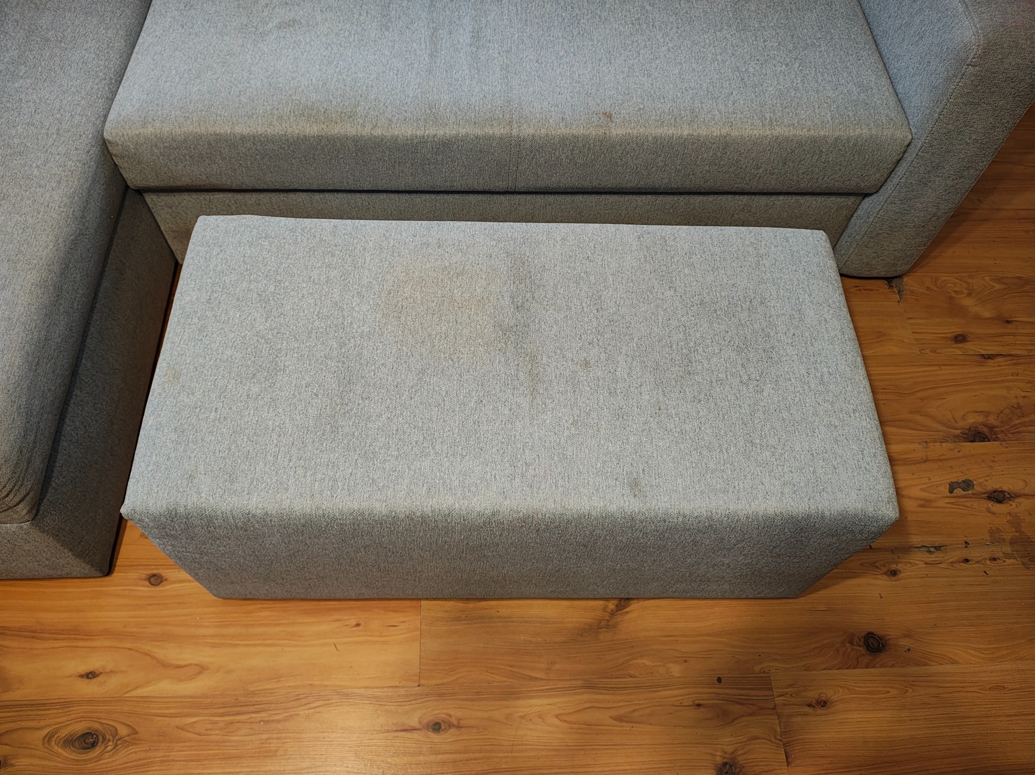 Szary puf z plamami na tle drewnianej podłogi i fragmentu sofy. Widoczne zabrudzenia na tapicerce przed czyszczeniem.