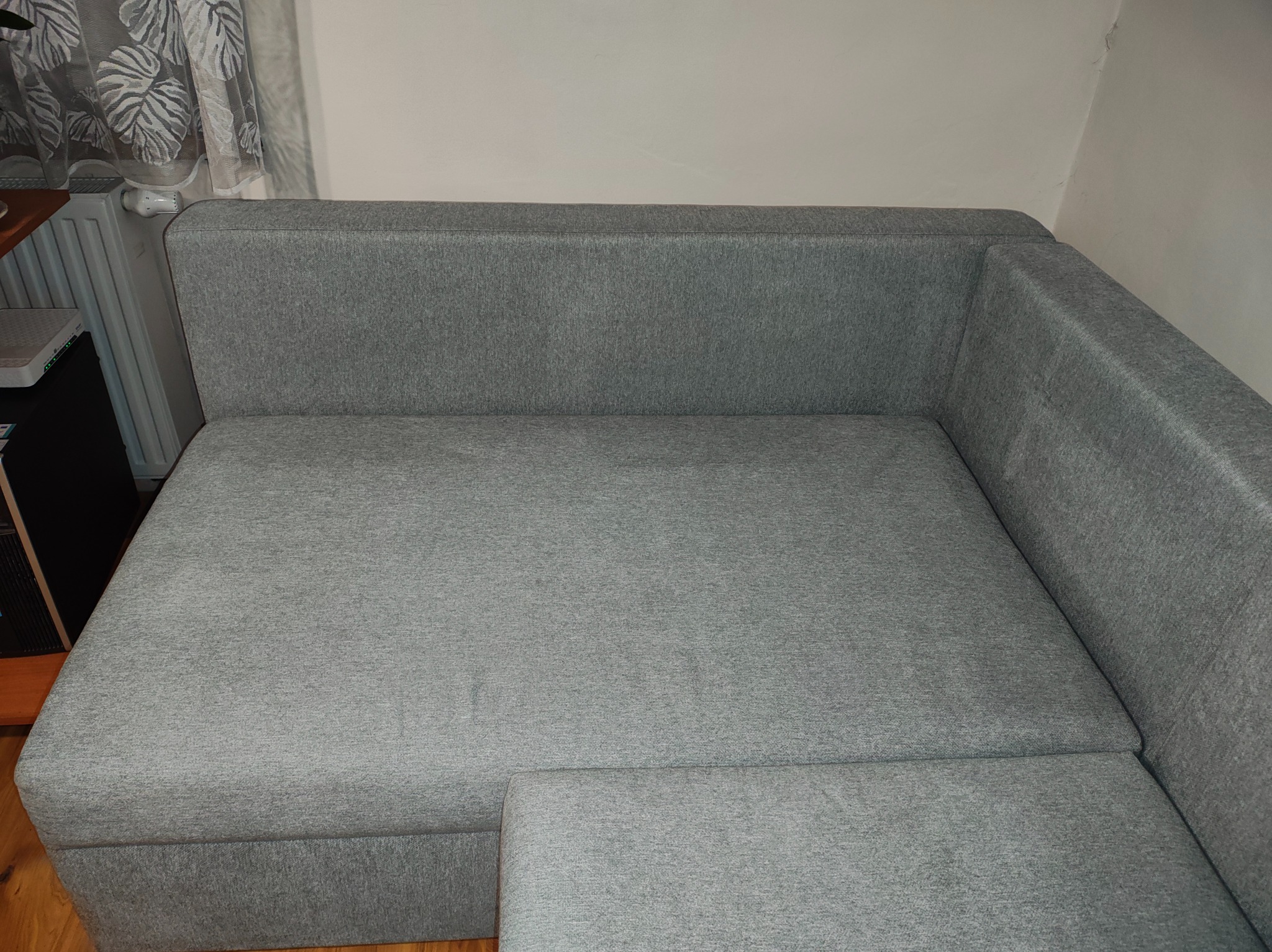 Szary narożnik z tkaniny, widok z góry, zarysowany kąt. Minimalistyczny design, jasne tło, widoczny fragment grzejnika i zasłony w tle.