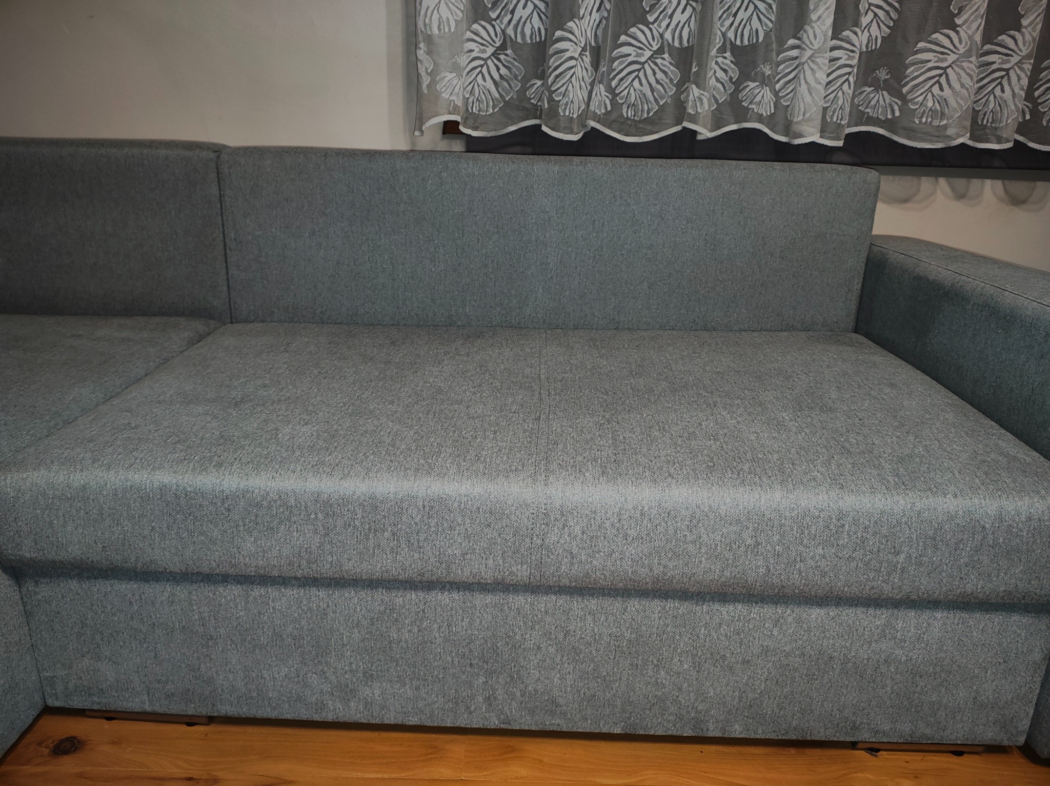 Szary narożnik z tkaniny w salonie, widok z przodu, z zasłoną w liście w tle. Widoczne drewniane panele podłogowe i fragment ściany. Minimalistyczny design.