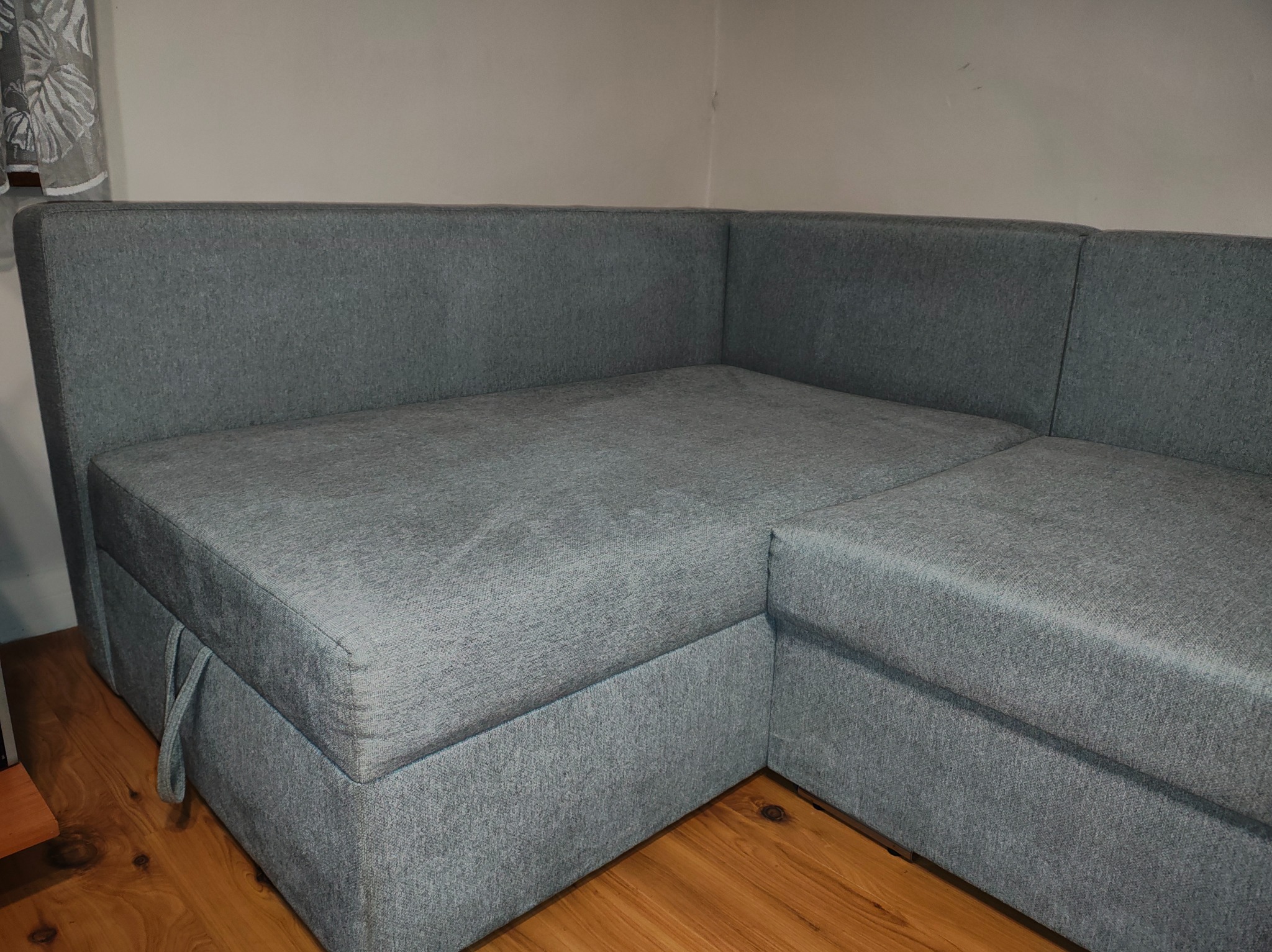 Szara, narożna kanapa tapicerowana w neutralnym odcieniu szarości, z widocznymi detalami szwów i fakturą materiału. Ujęcie z bliska, ukazujące narożnik mebla.