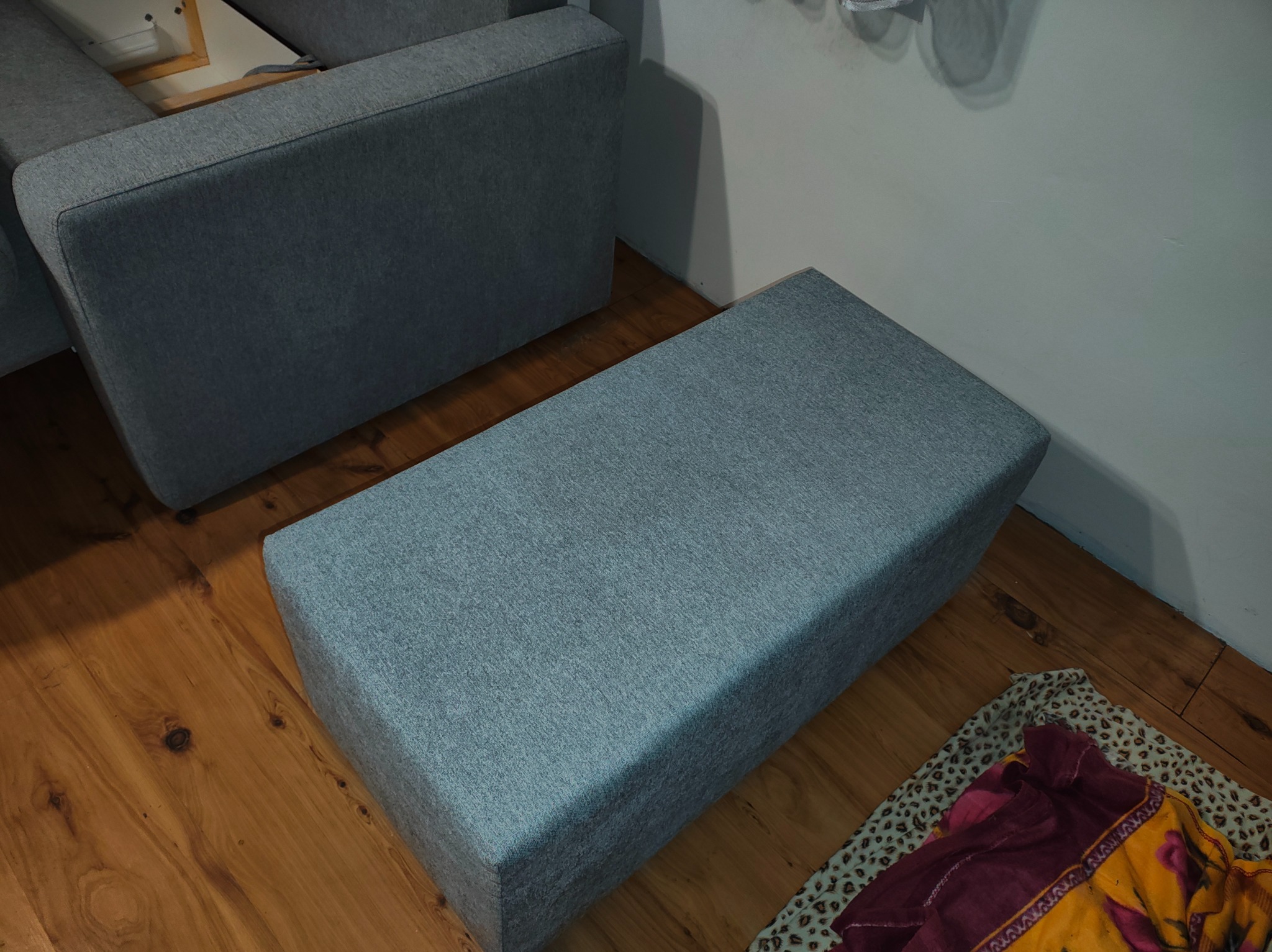 Szary puf tapicerowany obok sofy na drewnianej podłodze, z fragmentem wzorzystej tkaniny w rogu. Stonowana kolorystyka, proste kształty mebli.