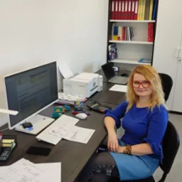 Kobieta w okularach siedzi przy biurku w biurze, na którym znajdują się dokumenty, komputer, drukarka i kalkulator.