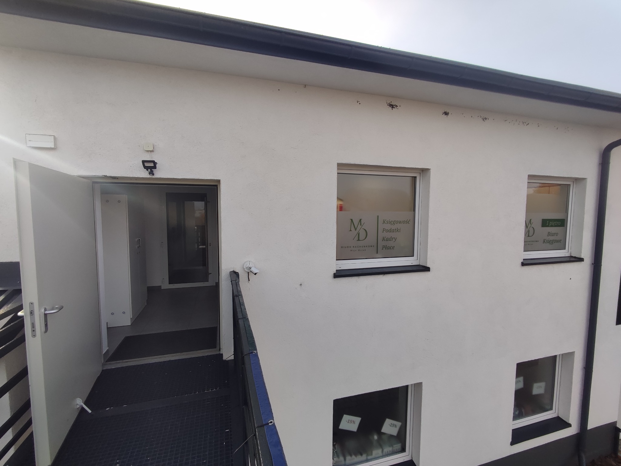 Wejście do biura rachunkowego z widocznymi oknami z naklejkami informacyjnymi o usługach: księgowość, podatki, kadry. Na oknach umieszczono również logo firmy i informację o promocji -15%.