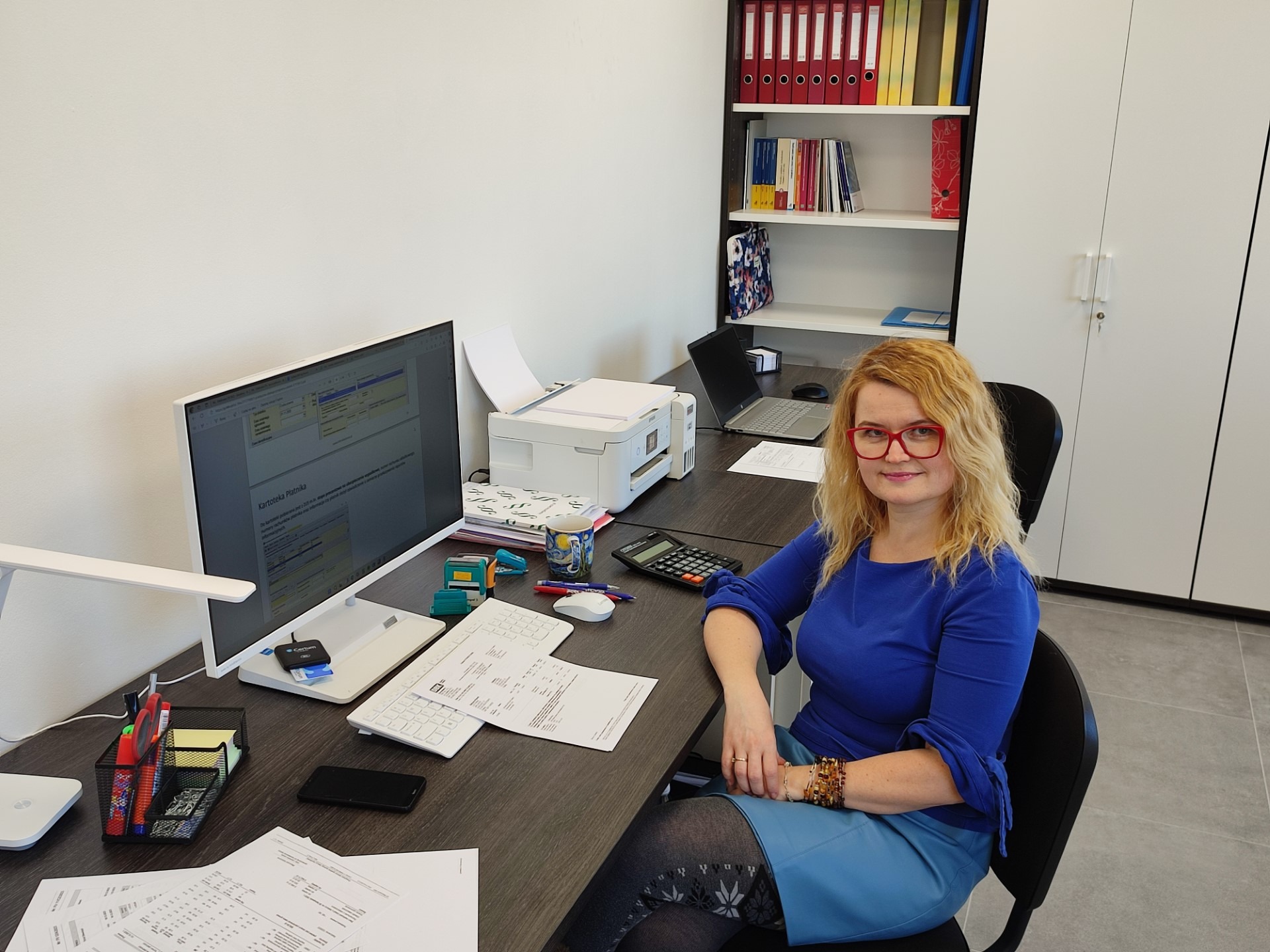 Kobieta w okularach siedzi przy biurku w biurze, na którym znajdują się dokumenty, komputer, drukarka i kalkulator.
