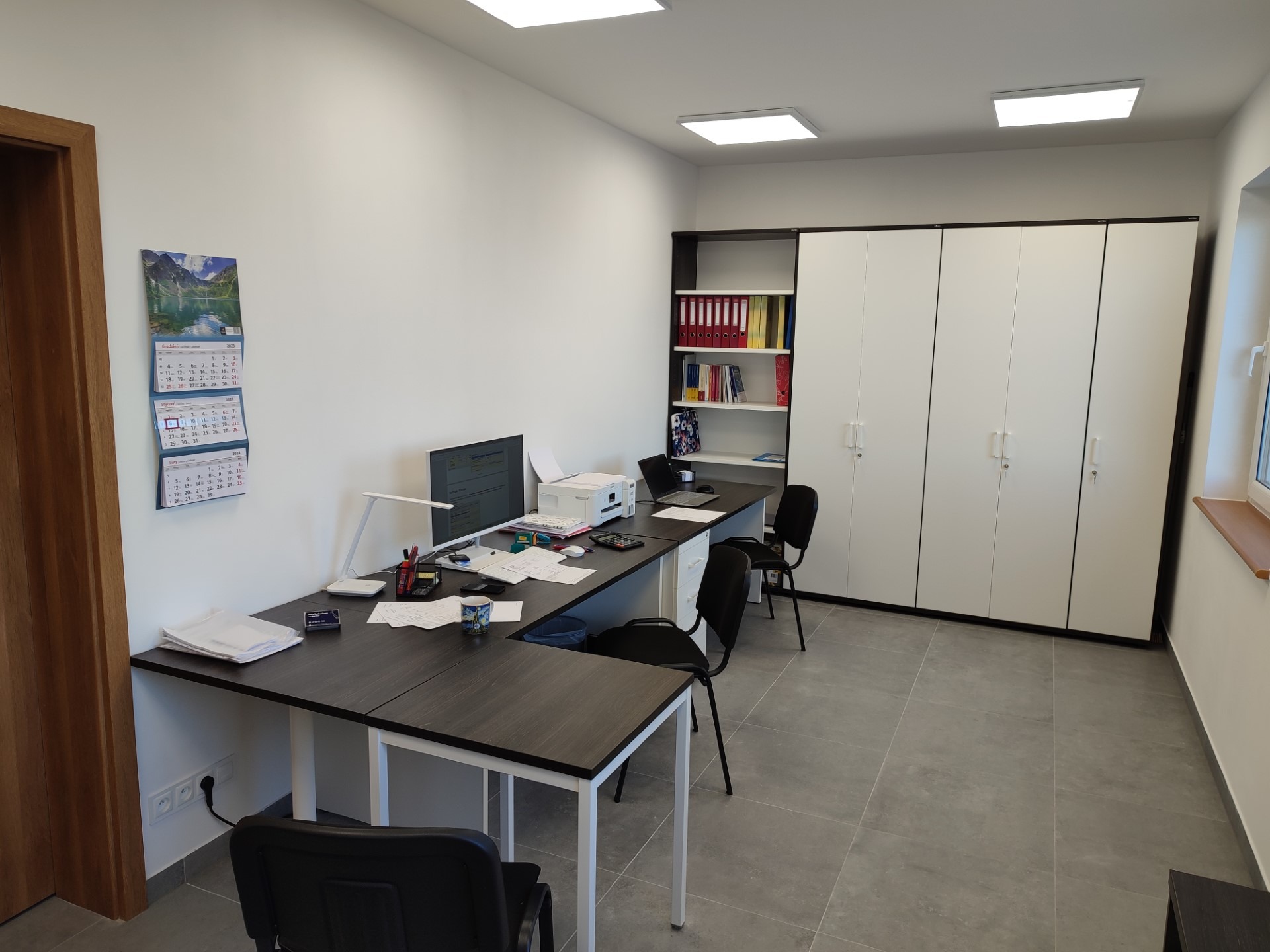 Nowoczesne biuro z dwoma stanowiskami pracy, komputerami, drukarką, kalendarzem na ścianie i szafą na dokumenty.