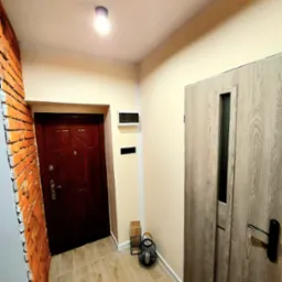 Korytarz po remoncie z brązowymi drzwiami wejściowymi, płytkami podłogowymi imitującymi drewno i fragmentem ściany z cegły.