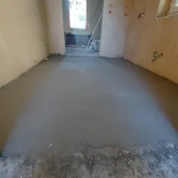 Wyrównana, szara wylewka podłogowa w trakcie schnięcia, widoczne niedoskonałości ścian i wystające przewody elektryczne, w tle okno z widokiem na zieleń.