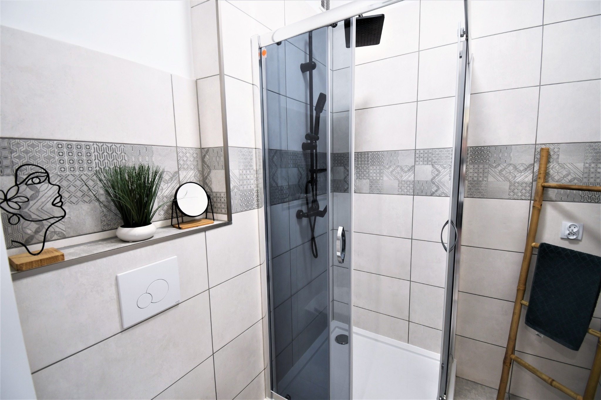Nowoczesna kabina prysznicowa z grafitową szybą, białe płytki z dekoracyjnym pasem, bambusowa drabina z ręcznikiem, minimalistyczne dodatki na półce.