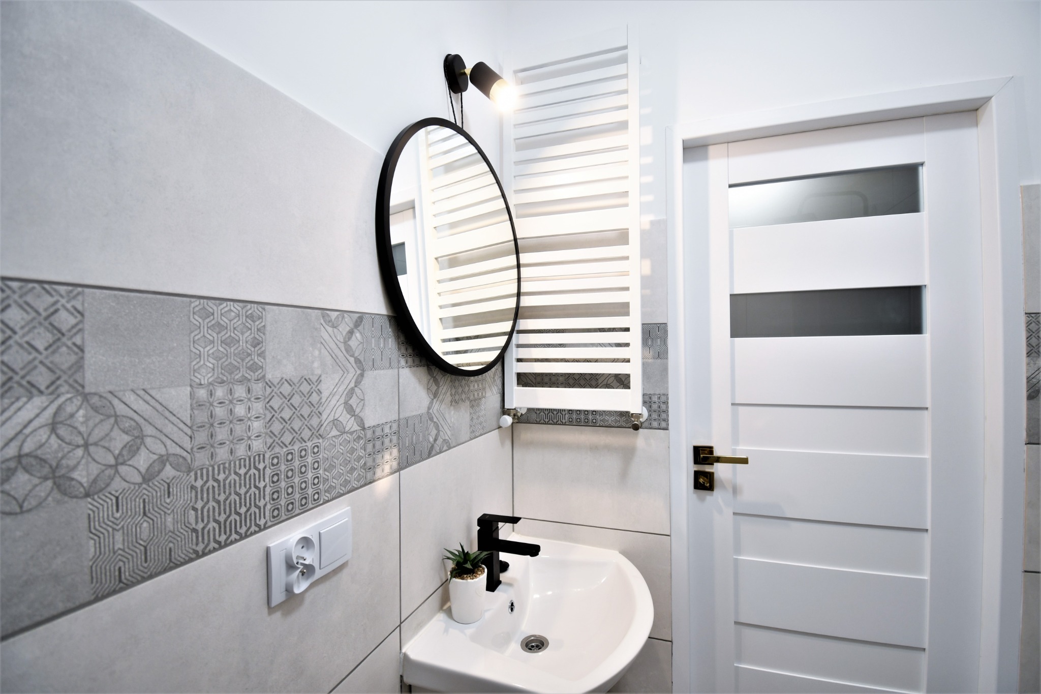 Nowoczesna łazienka z szaro-białymi płytkami dekoracyjnymi, okrągłym lustrem z czarną ramą, białą umywalką z czarną baterią i grzejnikiem drabinkowym.
