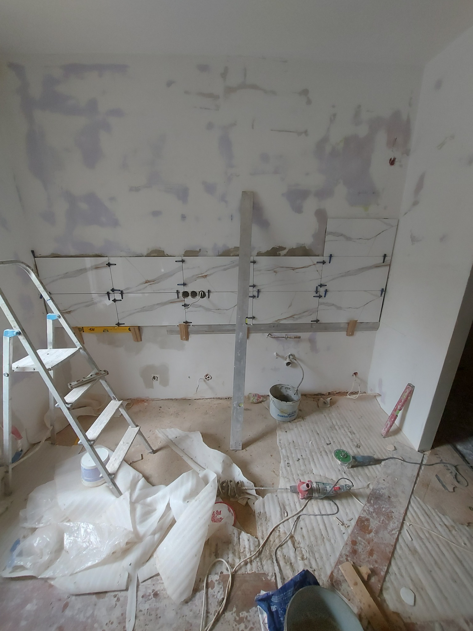 Ściana w trakcie układania płytek imitujących marmur, z widocznymi otworami na gniazdka, poziomica i drabina aluminiowa w tle, podłoga zabezpieczona kartonem.