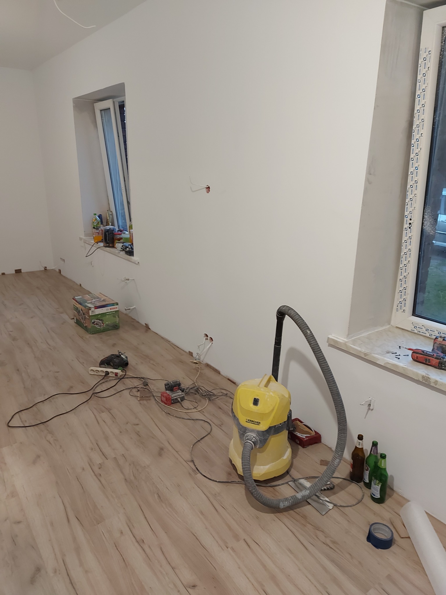 Wnętrze pokoju w trakcie prac wykończeniowych, widoczne panele podłogowe, ściany przygotowane do malowania, żółty odkurzacz przemysłowy, narzędzia i akcesoria budowlane rozłożone na podłodze...